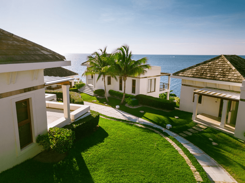 A modern multi-story luxury apartment or condominium building with white exterior walls and large balconies. The top floor features a glass-enclosed area. A long infinity pool is visible on one of the lower balconies, surrounded by wooden decking and outdoor furniture. The building is set against a backdrop of lush green tropical forest.