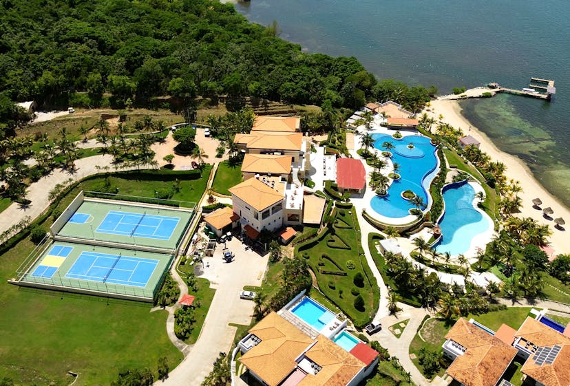 Aerial view of a luxury resort featuring two blue tennis courts, a large freeform swimming pool, and several buildings with terracotta roofs surrounded by lush greenery. The resort is located next to a body of water with a sandy beach and a dock extending into the water.