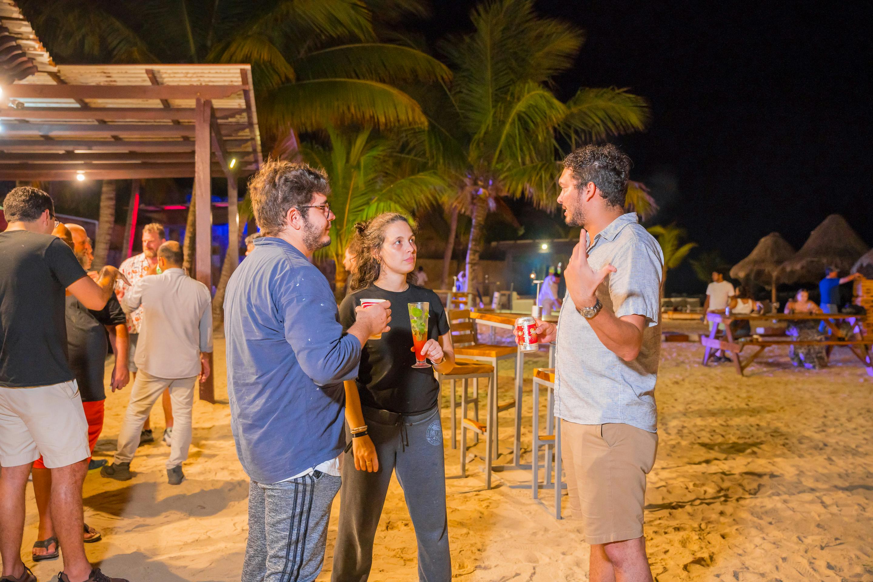 Group of people on a social gathering talking on a beach with drinks on their hands.