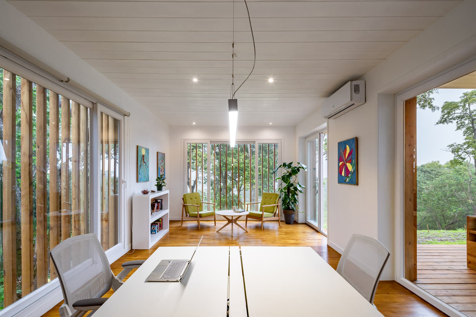 Bright and modern office space featuring a large table with a laptop, white walls, and wooden flooring. The room includes green chairs around a coffee table near a large window with vertical blinds, and colorful artwork on the walls, creating a welcoming and stylish environment.