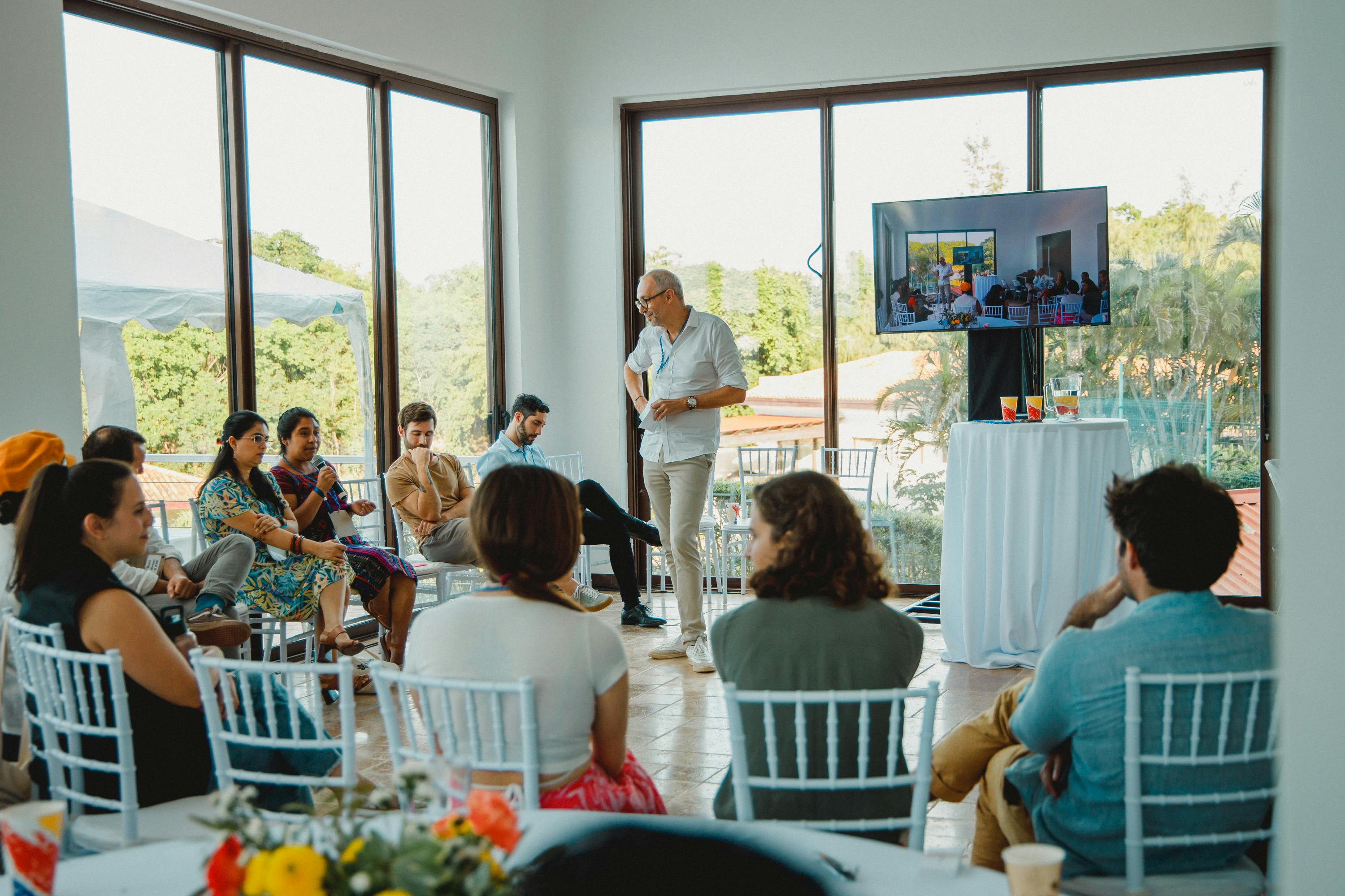 A group of people seated in a circle, participating in a discussion led by a standing speaker in a bright room with large windows. A television screen displays related content, and the room offers views of greenery outside, creating a relaxed and engaging atmosphere for the meeting.