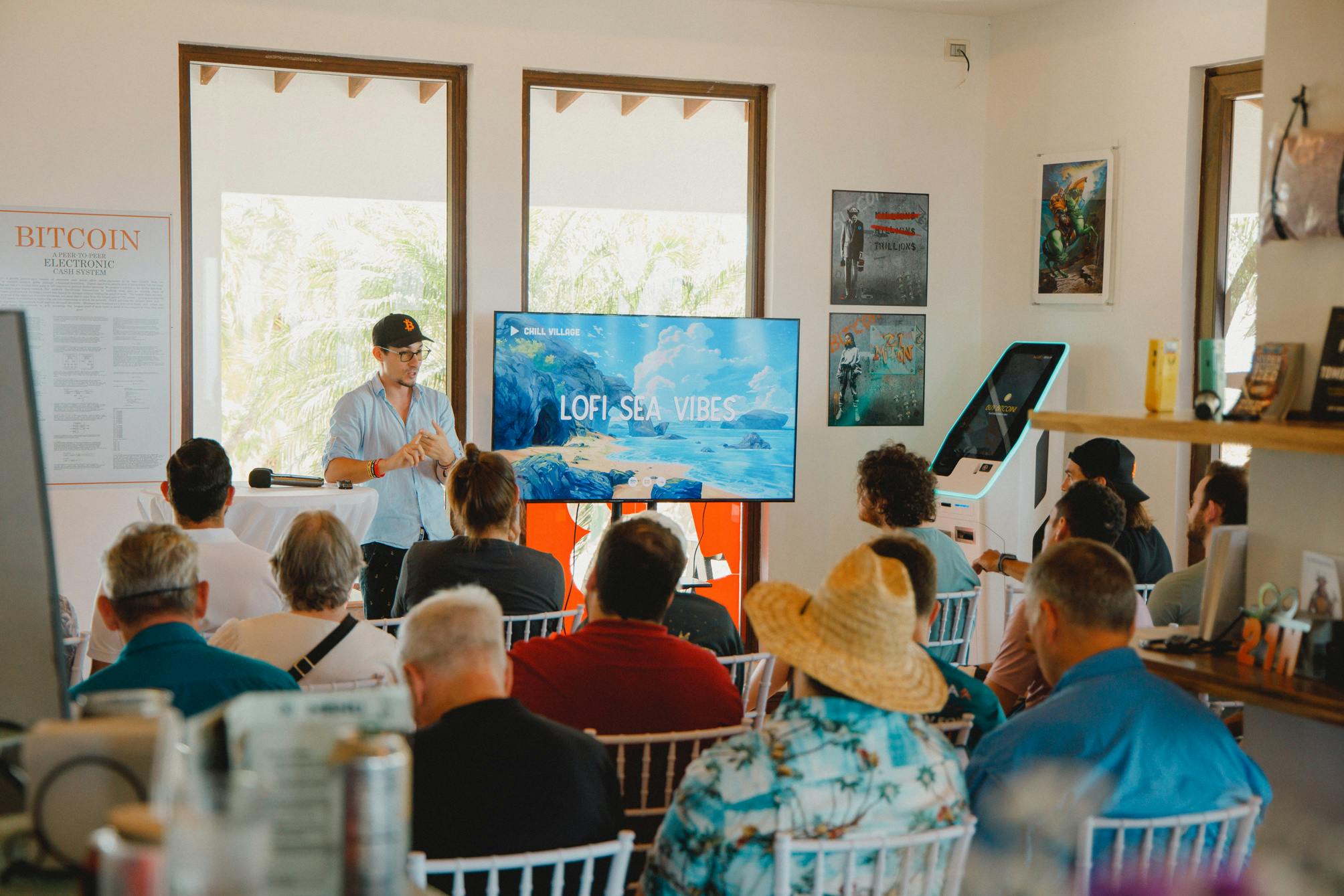 A group of people seated and listening to a speaker in a room, with a large screen displaying LOFI SEA VIBES. The setting includes posters on the walls and large windows providing natural light. The atmosphere appears casual and focused, with the audience engaged in the presentation.