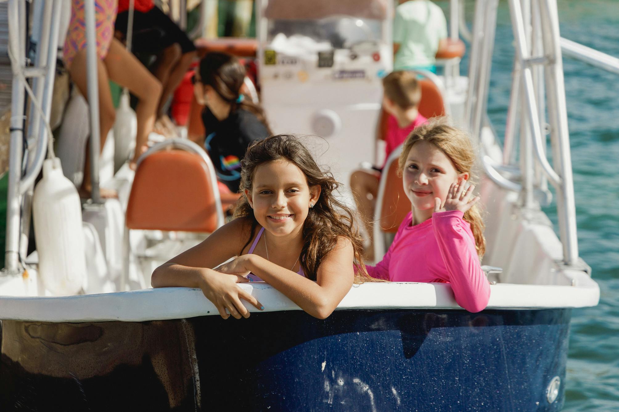 Two girls smiling and enjoying a boat ride, with one of them waving at the camera. Other passengers are visible in the background, creating a lively and cheerful atmosphere on the water.