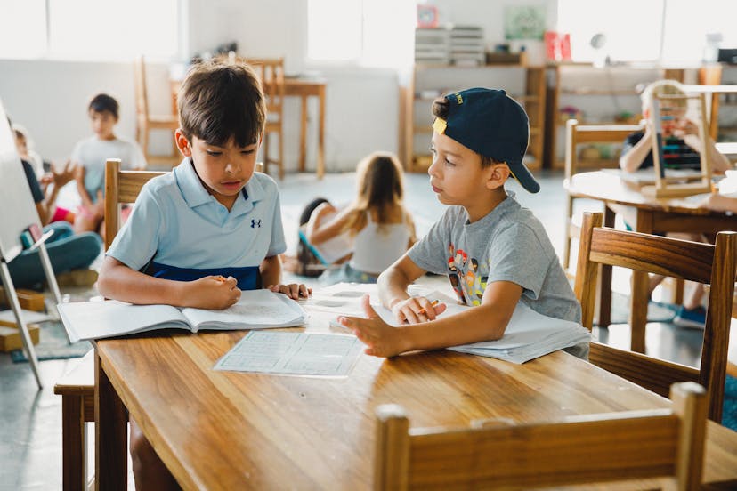 Two boys sitting at a wooden table in a classroom, engaged in a discussion over open books and papers. Other children are visible in the background, working in a collaborative and relaxed learning environment.