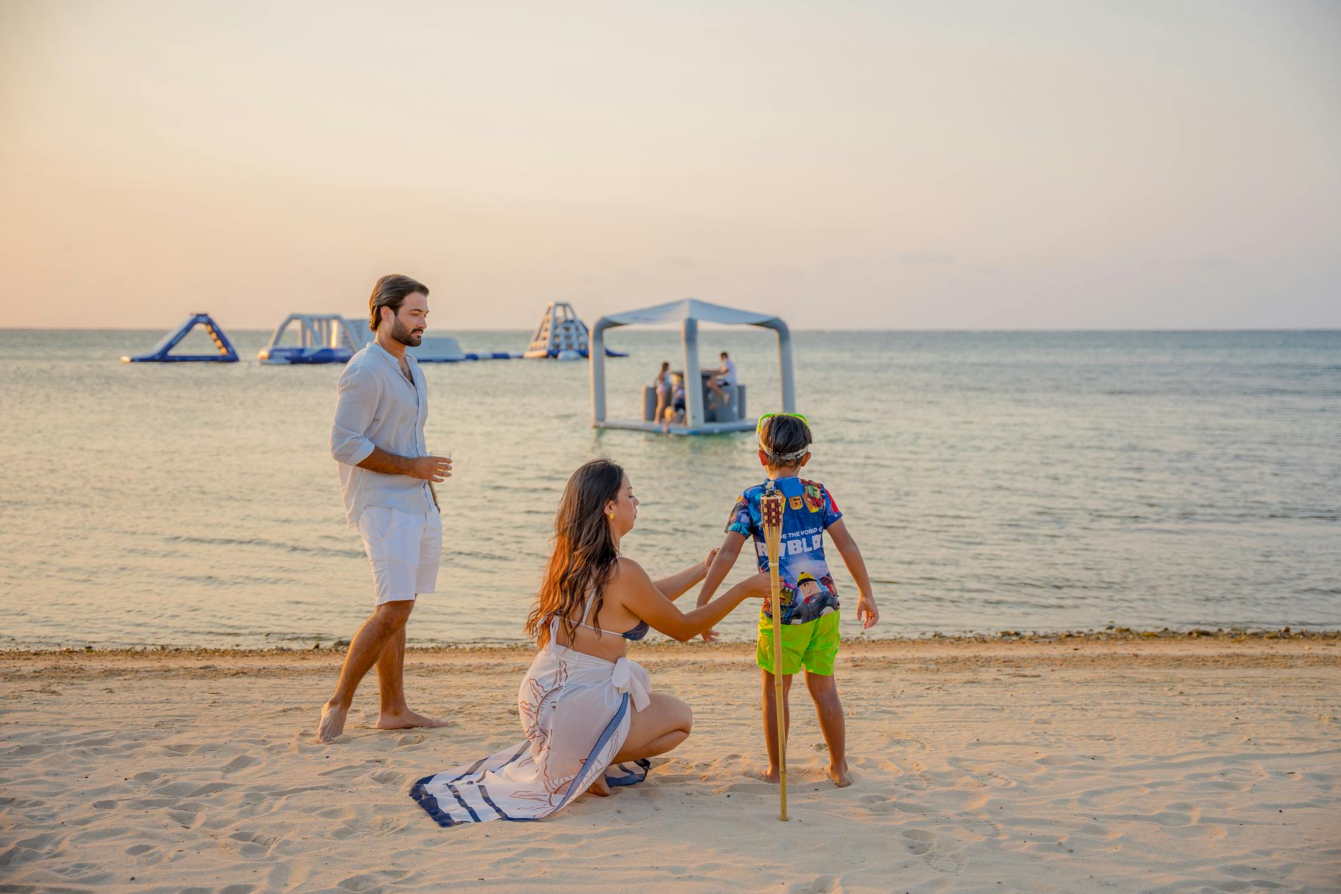 A father and a mom with a kid standing on a beach at sunset, with inflatable water structures floating in the background. The calm sea and pastel-colored sky create a peaceful and relaxed atmosphere, perfect for an evening by the water.