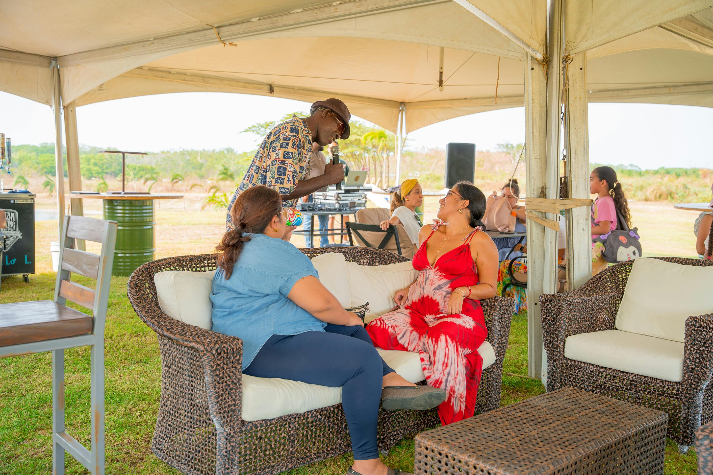 A group of people sitting on wicker furniture under a tent, engaging in a lively conversation. A man is speaking into a microphone, creating a relaxed and social atmosphere in an outdoor setting.