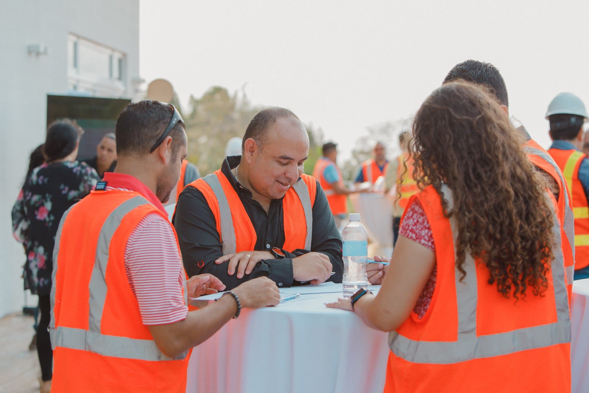 Un grupo de personas usando chalecos de seguridad naranjas, reunidas alrededor de una mesa y participando en una discusión o planificación. El entorno parece estar al aire libre, con algunos participantes sosteniendo documentos y otros interactuando en el fondo, sugiriendo un ambiente de trabajo colaborativo.