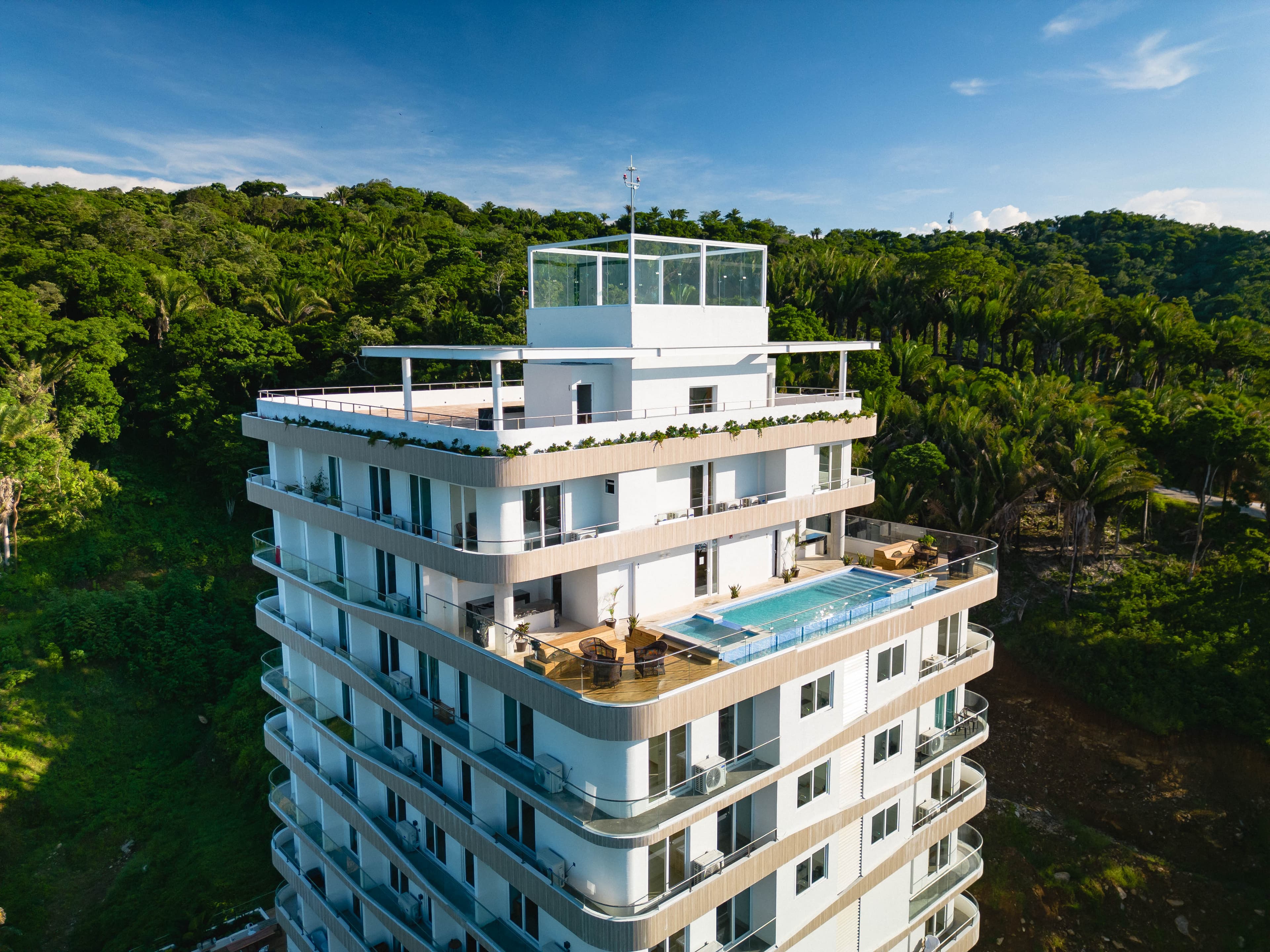 A tall, modern building under construction surrounded by dense greenery and palm trees, with the ocean visible in the background. The structure stands out against the blue sky with scattered clouds, indicating a coastal location with a mix of natural beauty and developing infrastructure.