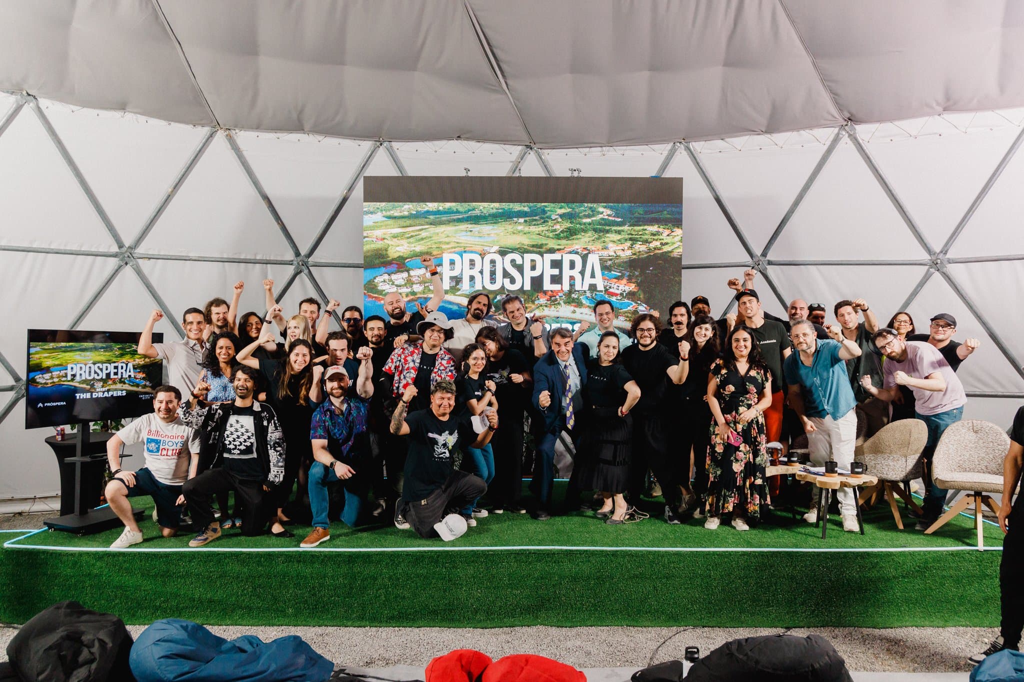 Una plataforma de madera con un letrero que dice PROSPERA, situada entre exuberante vegetación y árboles. La plataforma, elevada y rodeada de plantas y follaje vibrantes, crea un espacio exterior sereno y acogedor.