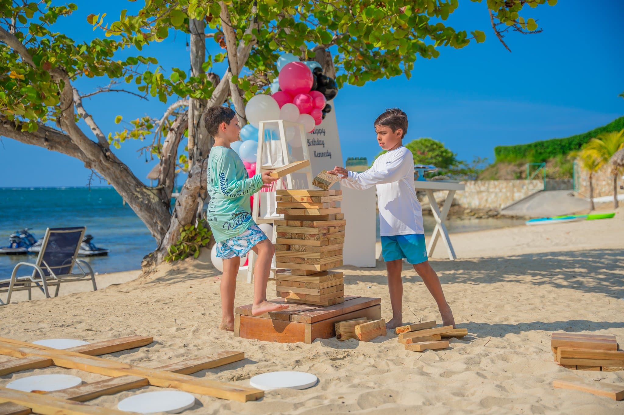 Two boys playing a game of giant Jenga on a sandy beach, with a backdrop of trees and balloons. The scene is bright and sunny, featuring a festive setup with colorful decorations and a beautiful ocean view in the background.