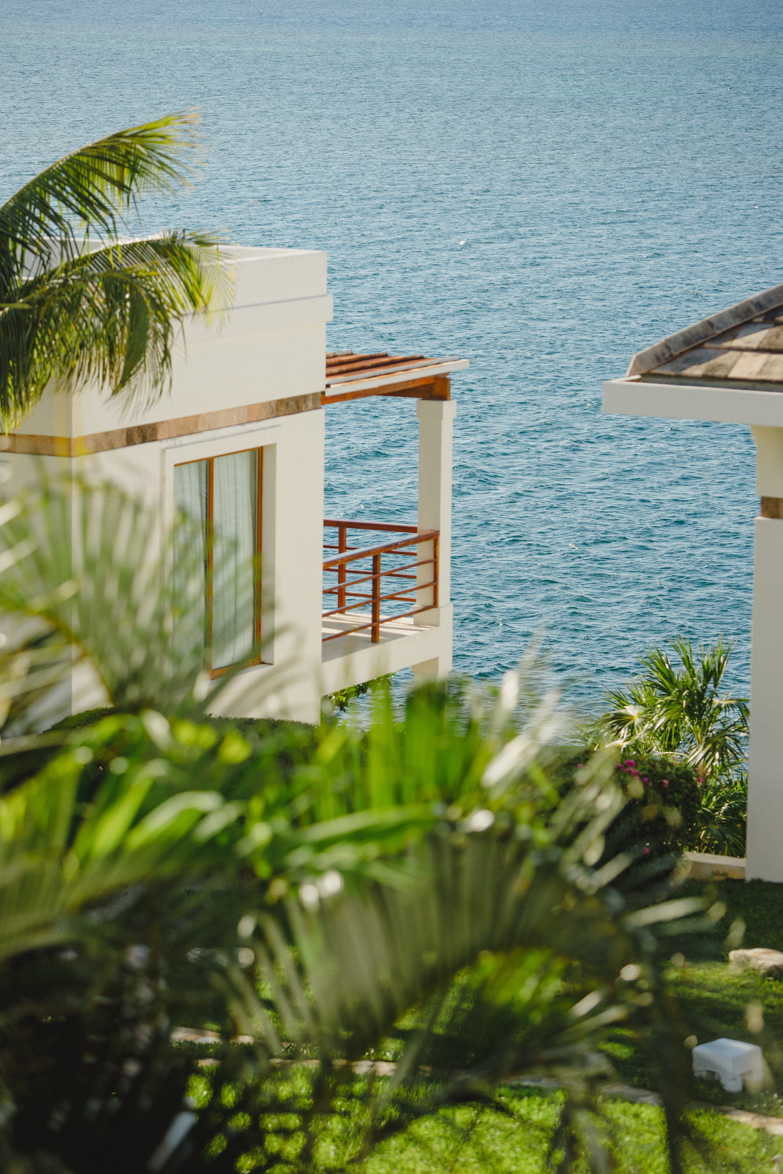 Aerial view of a waterfront resort featuring multiple two-story villas with tiled roofs, each surrounded by well-maintained lawns and palm trees. The villas are situated along a scenic coastline with calm waters and lush greenery, creating a tranquil and luxurious environment.